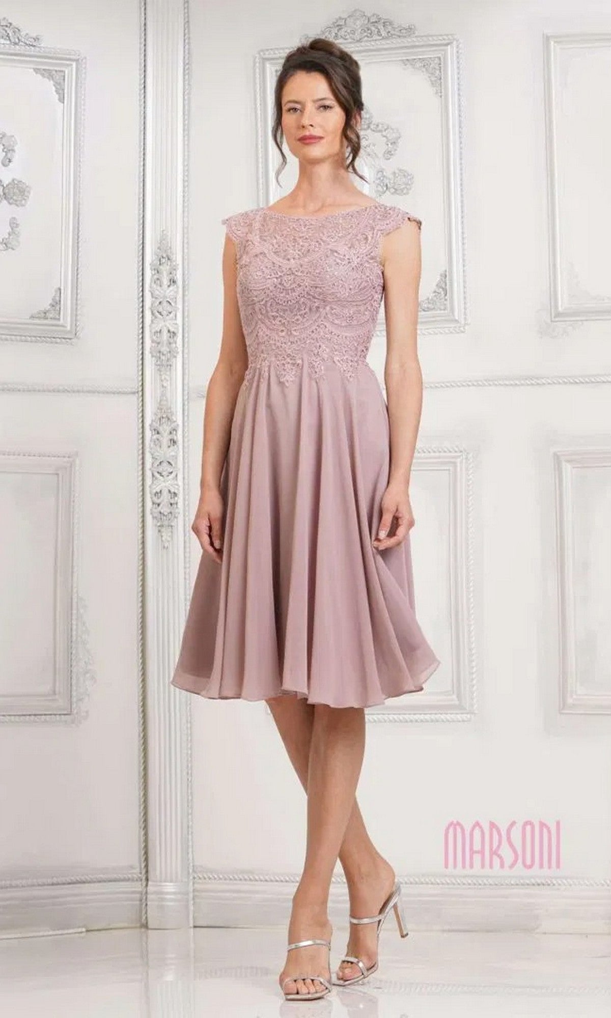 A woman stands indoors in a mauve Marsoni M238S knee-length semi-formal dress with a lace-embroidered bodice, chiffon skirt, cap sleeves, and paired with silver heels.